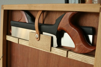 Le porte scie a ete une addition tardive. Il est en frene et en chene (ils viennent du bac a chutes). Le tasseau du milieu pivote grace a deux vis dont j'ai coupe la tete et qui servent d'axe. Ainsi ferme les scies ne bougent pas et ouvert les scies sont accessibles. Pour tenir la laniere de cuir j'ai recupere un bouton sur une vieille chemise :)